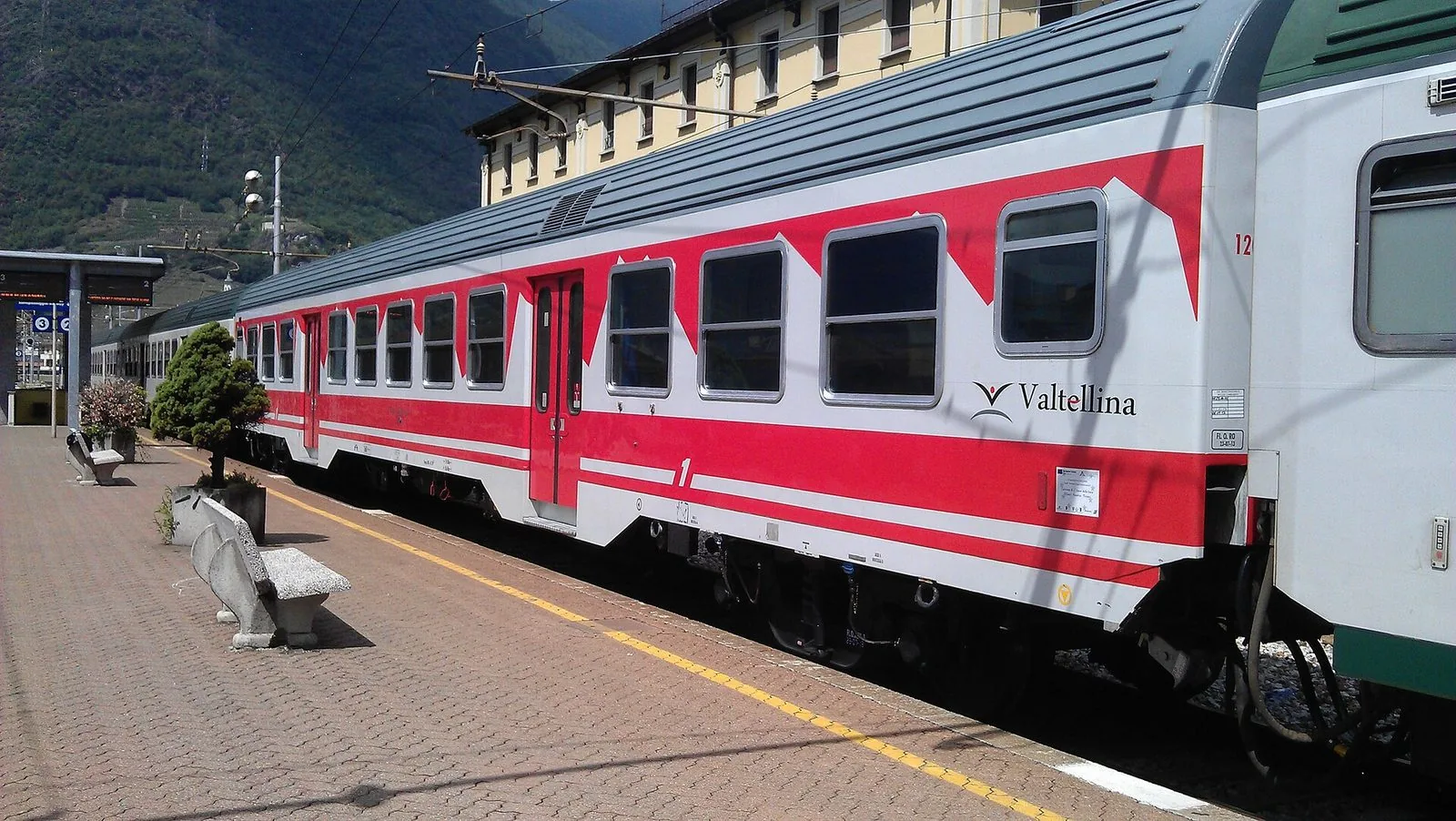 Carrozza ferroviaria con la livrea Valtellina ferma al binario di una stazione con le montagne sullo sfondo